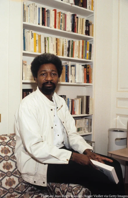 Wole Soyinka seated in front of book shelves. He is wearing a white jacket, white shirt and black pants. 