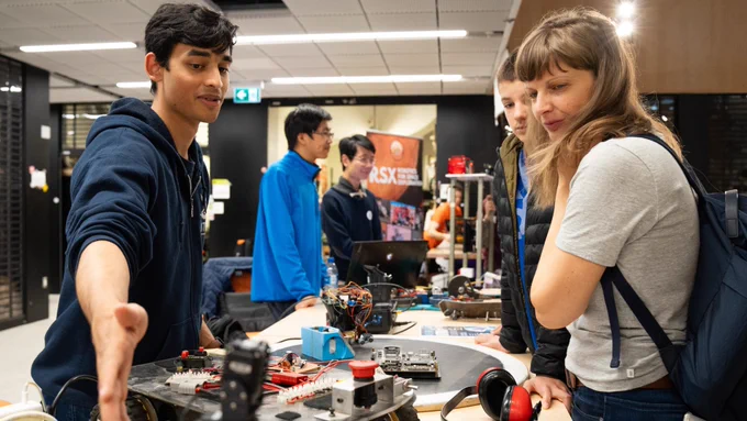 In the centre of the room sits a table piled high with robots and robot parts. On either side of the table, students and young individuals eagerly inspect the robots.
