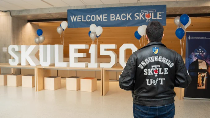 A person wearing an Engineering Skule™ jacket admires a blue banner that reads "Welcome Back Skule". Giant letters spelling "Skule 150" rest on the table surrounded by blue and white balloons.
