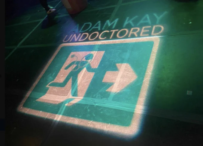 ADAM KAY - UNDOCTORED and a logo of a man with a stethoscope running through a door (like a fire exit sign) are projected in green onto the floor of a stage. It's called a Gobo, which Wikipedia tells me is an abbreviation of "Go-between". Not a very good abbreviation, if you ask me.