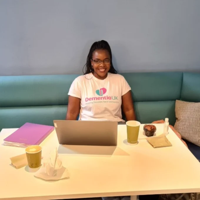 A woman called Tidziwe sits at a table. She is wearing glasses and a Dementia UK t-shirt. On the table is a laptop, takeaway coffee cups, a muffin and a notebook.