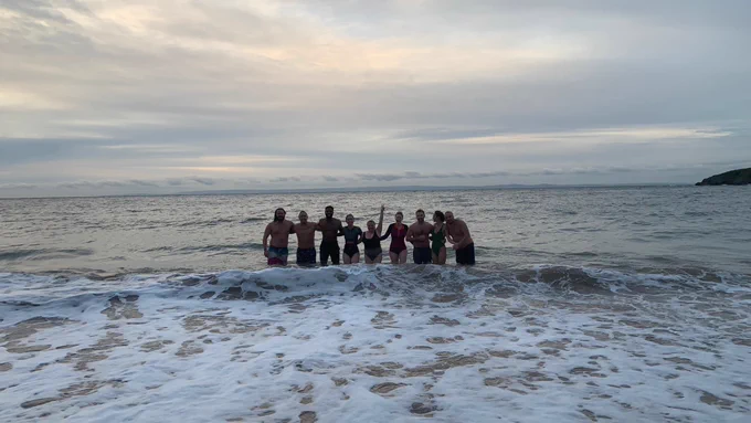 Photograph of group of people standing in waist deep cold water with one waving at the camera 