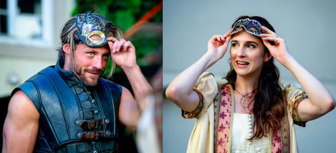 Taylor Napier as Romeo dressed in a dark blue/black leather period style vest with his huge arm muscles  lifting a masque ball mask and looking at Jessica Boone as Juliet lovingly. Jessica Boone is dressed in a cream period dress with long dark hair falling over her shoulder emailing and looking lovingly at Taylor as she also lifts a masque ball mask above her eyes
