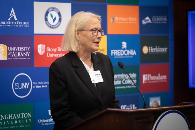 Woman with blonde hair, glasses and a black blazes standing at a podium with a banner i the background that contains many different logos from a variety of schools. 