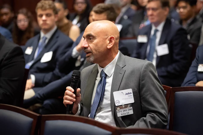 Man wearing a suit, holding a microphone in a crowd of people also wearing suits and sitting in chairs. 