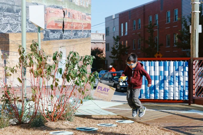 Image of a child at a playground jumping on circles on the ground with feet prints on them. A rectangular structure with blue and white movable pieces organized in rows is behind him. There is a building behind him that shows part of a large mural of and train.