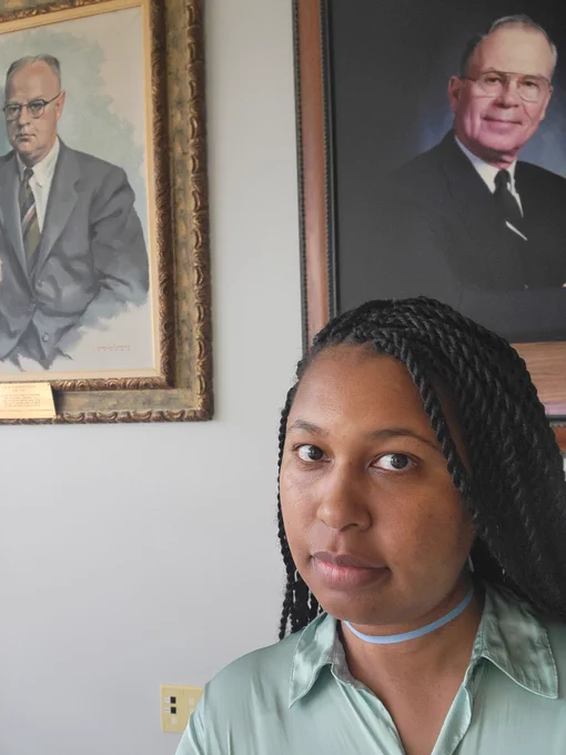 A Black woman with her hair in long twists takes a selfie in front of two portraits. The woman wears a satiny green button down shirt and has a N95 mask looped around her neck. The portraits depict two older white men, both of whom have gray hair and who are wearing glasses, one of whom is in a black suit and the other of whom is wearing a gray suit.