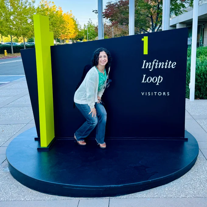 Me crouching in front of the 1 Infinite Loop sign at Apple. The sign has a large round back base that I’m standing on, a bright green number one that’s about 5 feet tall to the left of me, and white text on a black ground that says “1 Infinite Loop, Visitors”. I’m wearing gray jeans, a green top floral top and a tan sweater, smiling at the camera. 
