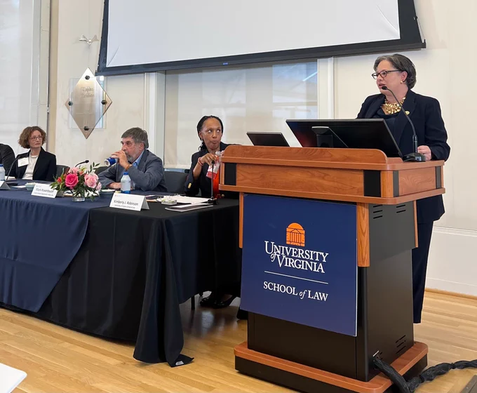Rep. McClellan delivers remarks behind a podium. On the podium is a navy blue and orange sign that reads "University of Virginia School of Law." Kimberly Jenkins Robinson, Mark Rosenbaum, and Kristine Bowman sit at a long table to the left of the podium. 