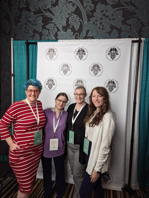 4 women, all pathology residents going into forensics, getting a group phone in front of the National Association of Medical Examiners banner at a conference. Everyone is well dressed and smiling.