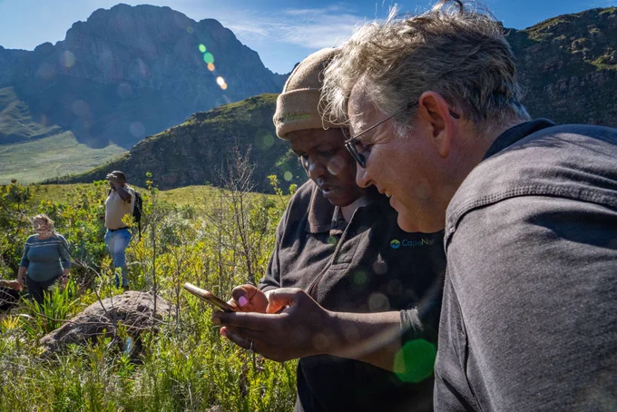 Photo credits: Jeremy Shelton

Alt text: Two people looking at a phone. They are standing in a grassy area with mountains behind them. There are two more people standing to the left of them looking out into the field.