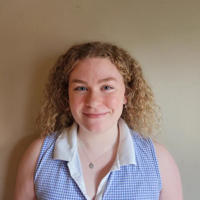 Image of a young woman smiling. Woman has curly blond hair and is wearing a blue and white checkered shirt.