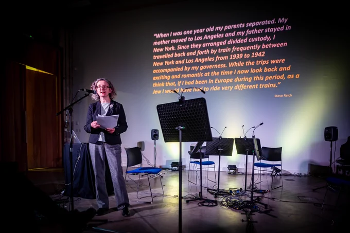 A photo of Dr Morag Grant introducing the Steve Reich: Different Trains concert, text from Steve Reich is projected behind her