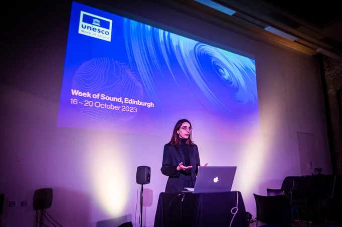 A photo of Dr Eleni-Ira Panourgia giving the welcome talk at the event, with the UNESCO Week of Sound projected behind her