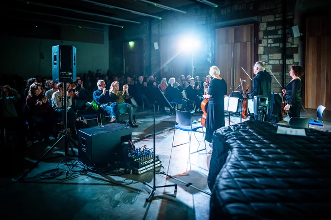 A photo from the back of the stage, with performers Jeff Moore (Violin), Ariane Todes (Violin), Charles Mutter (Viola), Dee Isaacs (Cello) on the right, and the audience clapping on the left
