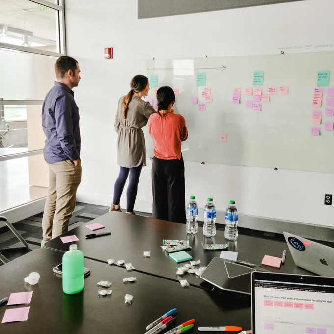 Kelli Lucass, co-founder and Chief Design Officer of Lunarlab, collaborating with clients on a whiteboard covered in post-it notes. The table is adorned with a mix of mints, water bottles, sharpies, and laptops.