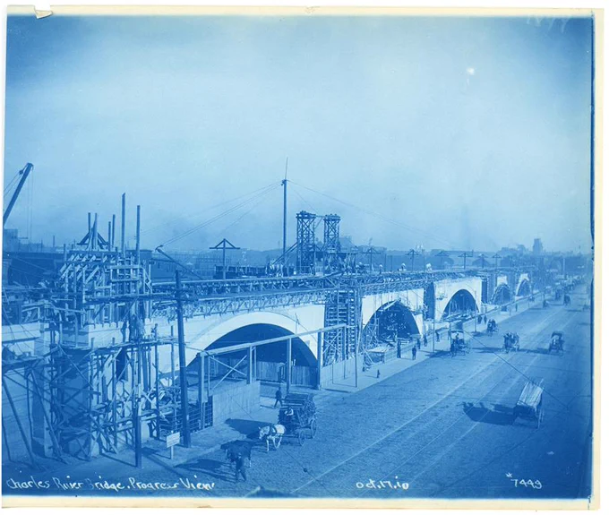 Cyanotype photograph of construction on the Charles River Bridge. Bridge is covered in scaffolding and horse drawn carts are on the road below.