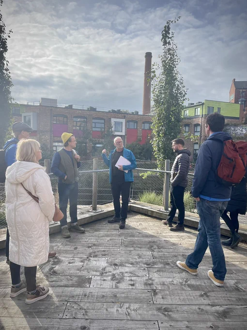 A group of people on a tour gathered around a man guiding them and pointing at a reused building in the background.
