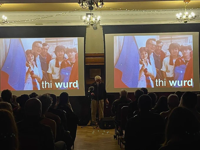 Colour image of Bernard MacLaverty standing at a microphone, between two screens, the backs of the audience’s heads in semi-darkness watching him.