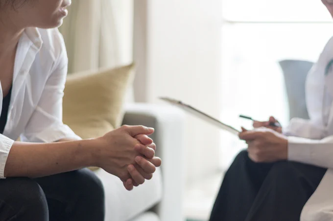 A doctor holding a clipboard and pen sits on a couch opposite a woman.