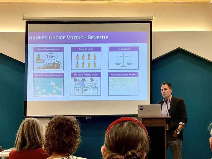 White man at a podium next to a slide illustrating the benefits of Ranked Choice Voting.