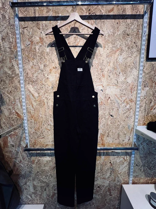 A pair of black dungarees hung on a clothes hanger against a wooden backdrop. 