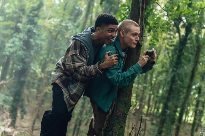 Charlie (played by Jay Lycurgo) and Steff (played by Lewis Gribben) are in a well-lit forest. Charlie is holding onto Steff who is taking a picture and they're both happy. 