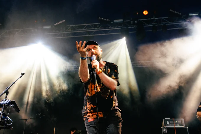 Pictish Trail sings into a microphone with his arm raised in the air, on stage at Green Man 2023. 