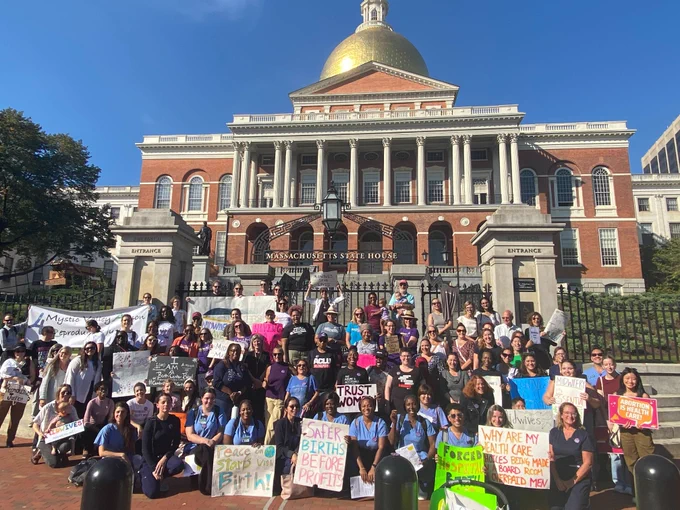 The Rally for Midwives stands in front of the State House for a group photo. Many attendees have signs. 