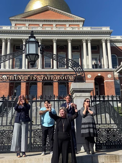 Rep. Decker, wearing all black, stands in front of the State House with her fists raised. Four representatives stand behind her. 
