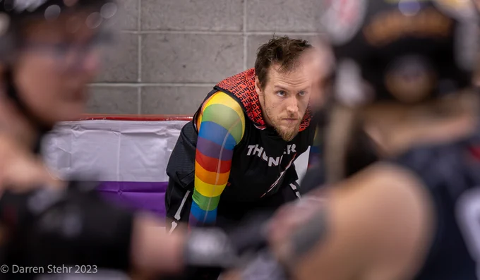 A person in a Thunder hoodie with a rainbow mesh top underneath is staring intently through a crowd of derby skaters