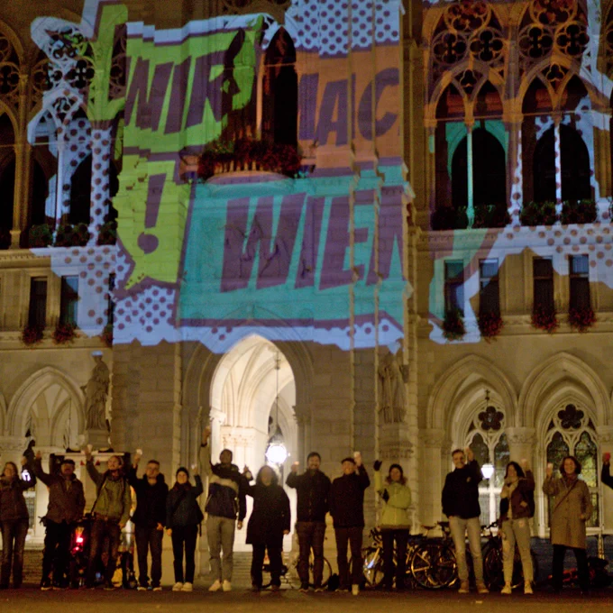 #WirMachenWien Aktivist*innen stoßen vor dem Rathaus mit Sekt an. Dahinter ist das #WirMachenWien Logo auf da Rathaus projeziert.