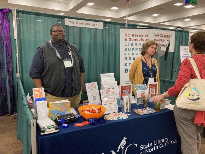 Two State Library staff members standing behind a display table at the NC Library Association Annual Conference. A visitor to the table is talking to them. 