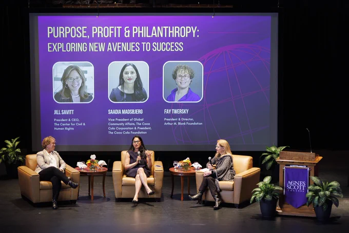 A group of professionals sit in comfy armchairs on a stage amidst conversation.