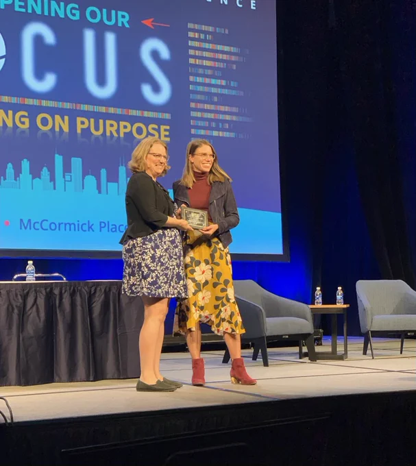 Two women pose on stage at a conference holding an award plaque between them.