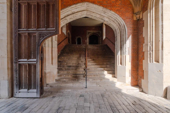Photograph looking towards a stone staircase, which is through a stone archway in a brickwall. There is a wooden handrail up the centre and arched windows and doorway can be seen at the top of the stairs. In the foreground is a cobbled floor, sunlight is coming in from the right and to the left is a wooden carved door with an arch cut from the lower right section.