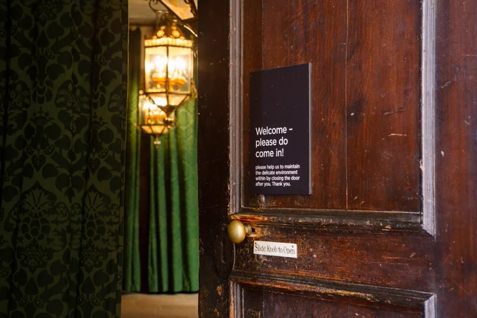 Photograph of a partially open old wooden door to the right with a shiny brass doorhandle with wood grain visible in patches where the darker brown stain has been worn away. There is a sign saying 'Slide Knob to Open' right by the knob and another sign above saying 'Welcome - please do come in! please help us to maintain the delicate environment within by closing the door after you. Thank you.' To the left of the frame you can see into the room, with a patterned green curtain in the foreground and distance, with two large brass and glass lanterns hanging from the ceiling.