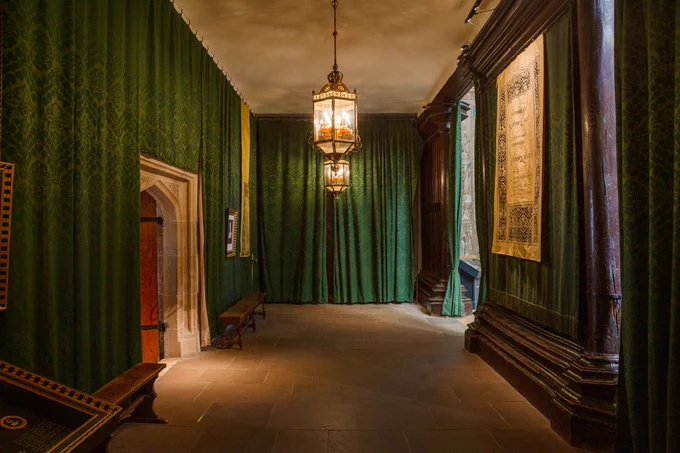 Photograph in Hampton Court Palace showing a dark room lit by two ornate brass and glass lanterns hanging from the ceiling in the middle, stone floor, the walls lined with patterned green curtains, a stone arched doorway to the left, wooden benches along the lefthand wall, a wooden wall to the right with a tapestry hanging in the centre and an opening at the far end into a daylit room.