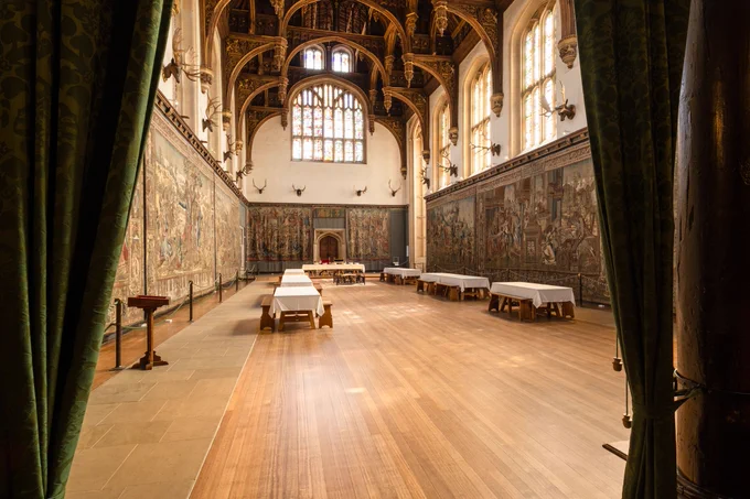 Photograph in Hampton Court Palace looking through two green curtains that are tied back either side of the frame, into a large room with wooden floor, tapestries and stag heads lining the walls, banquet tables with white table cloths and wooden stools around the edges, ornate carved wooden ceiling and an arched doorway at the far end.