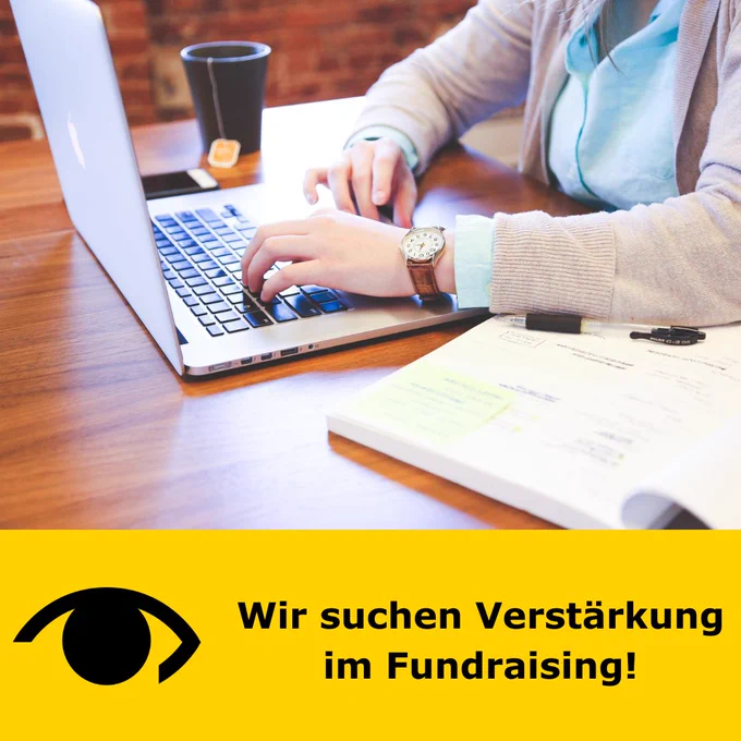 Auf einem Holztisch steht ein aufgeklappter Laptop vor dem ein Mensch stitzt, aber es sind nur die Arme und ansatzweise der Oberkörper zu sehen. Neben dem Laptop liegen eine Tasse Tee und Unterlagen. Darunter ist ein gelber Balken mit dem schwarzen Logo der PRO RETINA und daneben steht: Wir suchen Verstärkung im Fundraising!