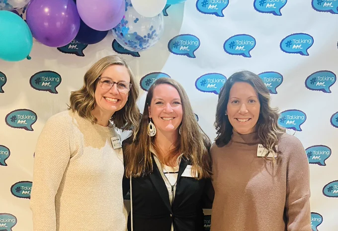 Three women standing in front of a back drop that says #TalkingAAC. A blonde woman on the left is wearing glasses, a white sweater, and smiling. A brown haired woman wearing a black shirt is smiling and standing in the middle. A brown haired woman wearing a brown sweater is on the right and is smiling.