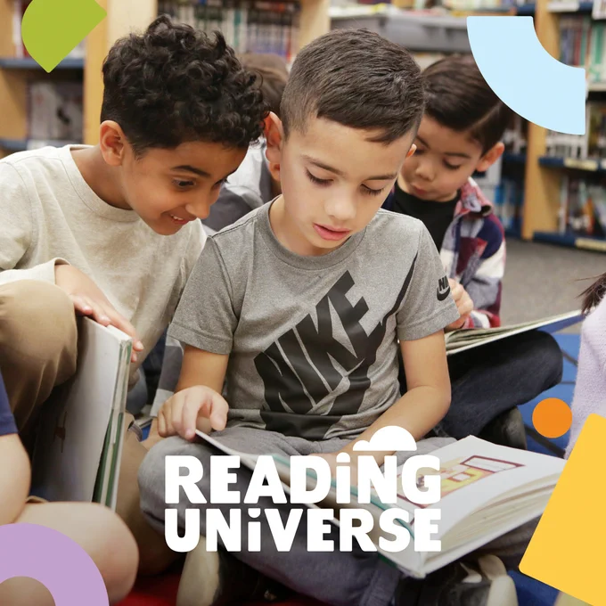 One child reads a book, while another looks over their shoulder excited about what's in the book.