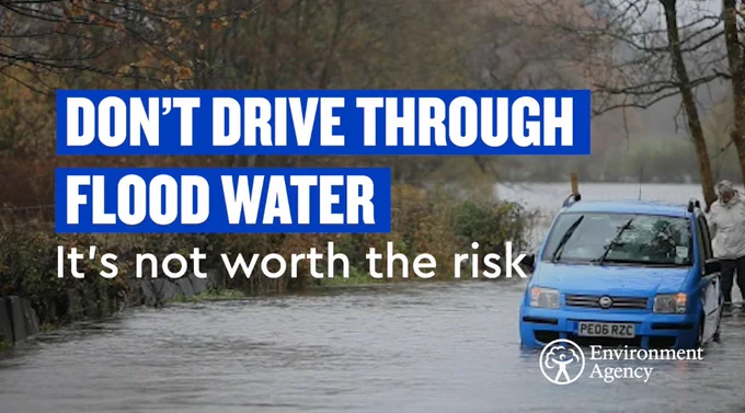 A blue car is seen stuck in floodwater up to the number plate. The road has become a river. A man stands behind the car. In white capital letters, the graphic says DON'T DRIVE THROUGH FLOODWATER. Below it in sentence case: It's not worth the risk. A white Environment Agency logo is in the bottom right-hand corner.  
