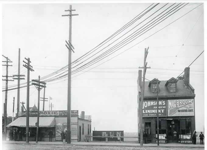 street scene with wooden buildings, overhead wires, and advertisements for Moxie, Johnson's Linement, and Nu-Broom