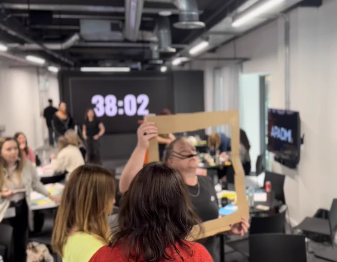 A person holds up a cardboard frame using pipe cleaners as a moustache to act out a scene to the rest of their team
