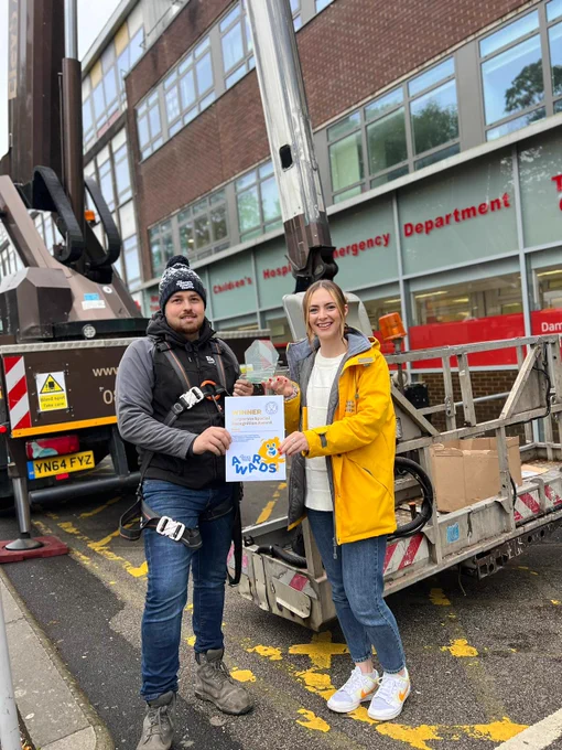 A lady in a yellow jacket hands a certificate and a trophy to a man wearing winter clothes and a harness. There is an elevated platform behind them with boxes on it. 