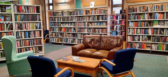 Bookshelves full of books in Orkney Library & Archive. At the front is a coffee table with a sofa and chairs around it.