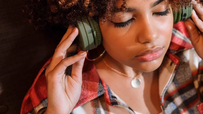 A woman, shown from the shoulders up, holds a pair of over ear headphones to her ears and has her eyes closed. 
