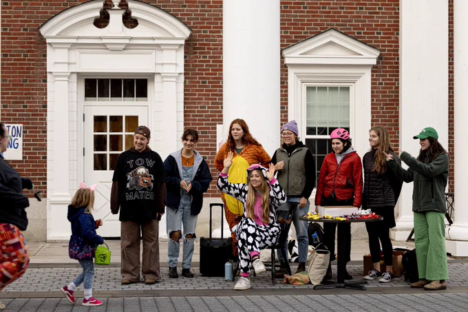 A group of college students in various states of costumed dress, stand and welcome a group of trick-or-treaters along Alumni Walk.