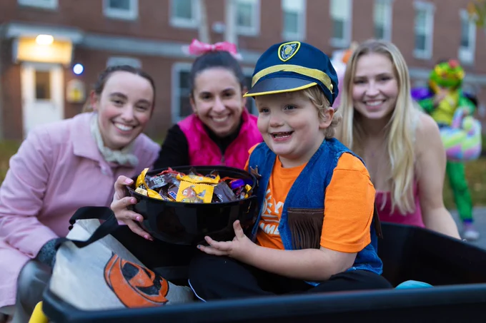 Young boy smiles while holding large bowl filled with candy. Three female college students smile behind him.